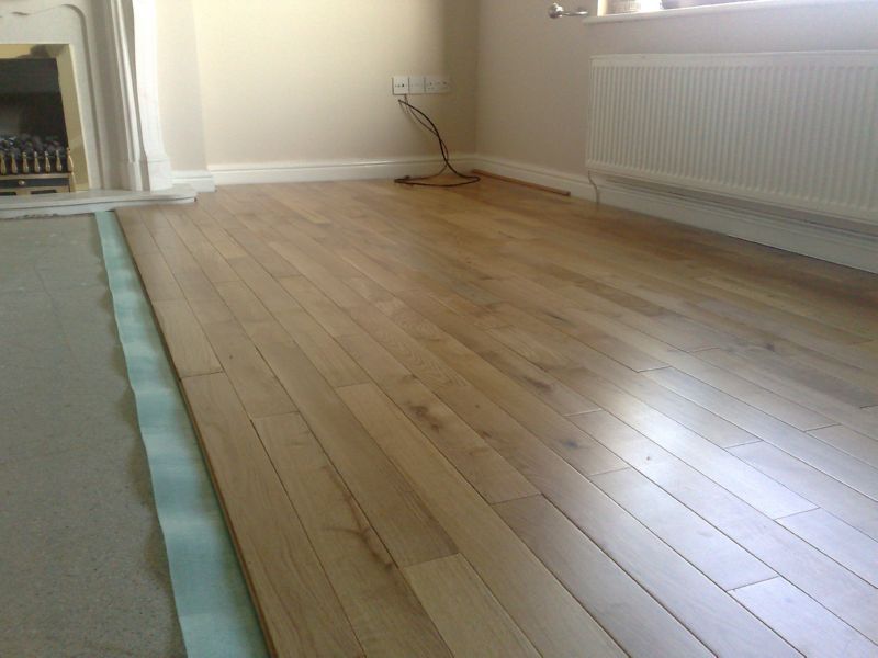 Układanie jasnej, drewnianej podłogi w pokoju z kominkiem i grzejnikiem; widoczne przygotowane podłoże z folią izolacyjną.